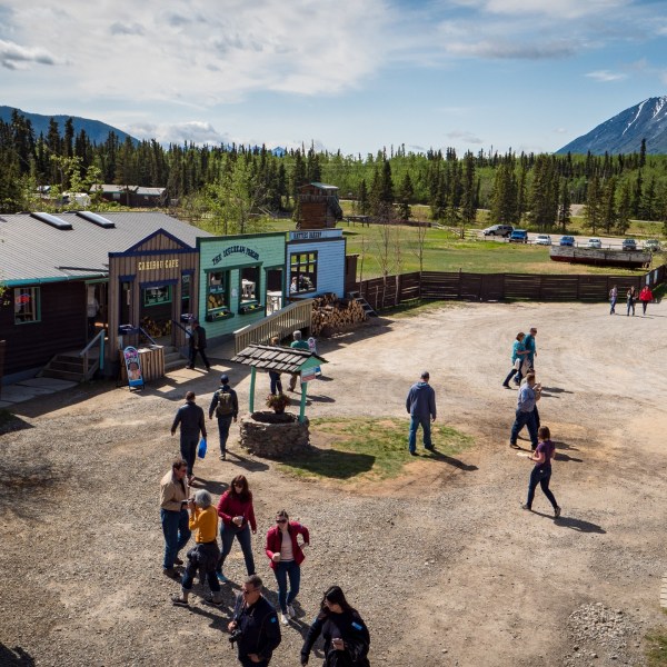An elevated wide shot of a rustic tourist outpost with several wooden buildings, a dirt courtyard filled with people, and snow-capped mountains in the background.
