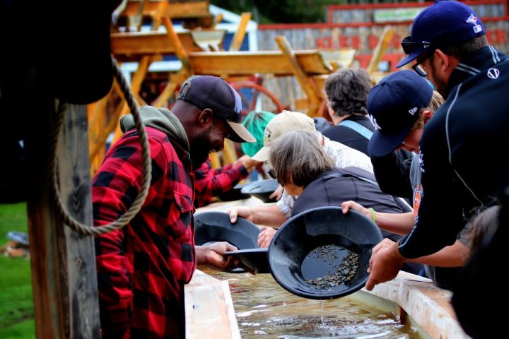 A man in a red and black plaid jacket smiles while showing a group of tourists how to pan for gold in a long wooden water trough.