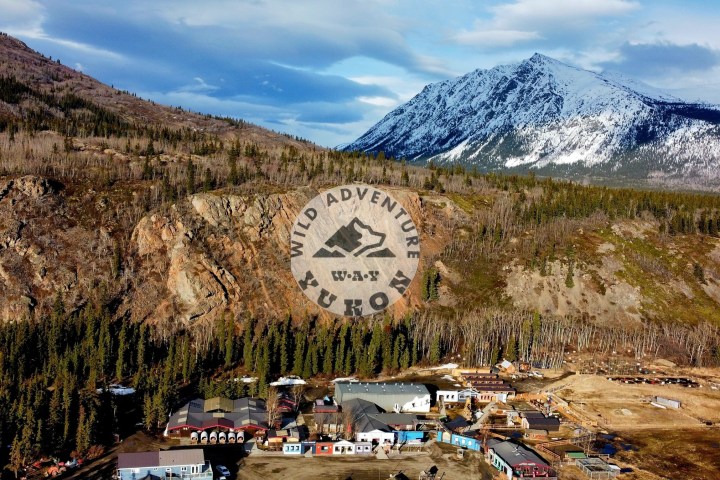 Aerial view of buildings in a forested mountain valley with snowy peaks and 'Wild Adventure Yukon' logo overlay.