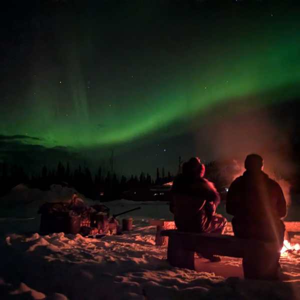 Two people by a campfire under the northern lights in a snowy landscape.