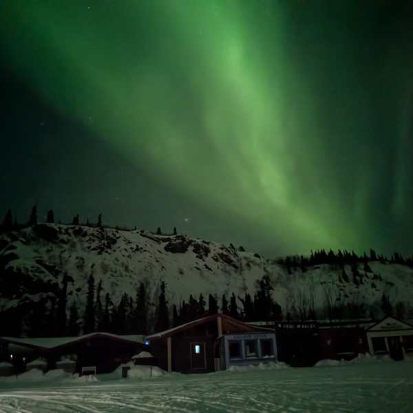 Northern lights over snowy landscape with cabins and trees.